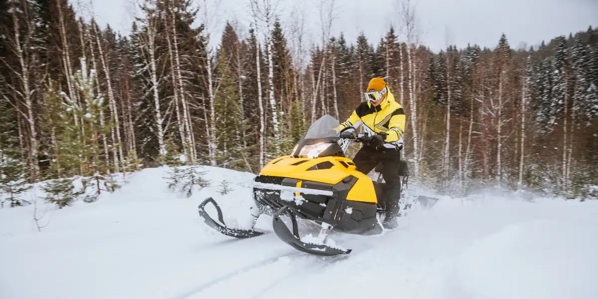 A man rides a yellow snowmobile through a winter forest