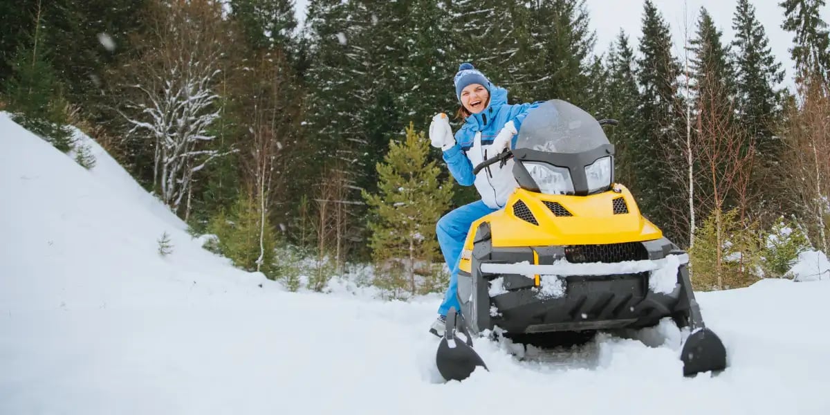 A woman is sitting on a yellow snowmobile in a winter forest drinking from thermostat and laughs